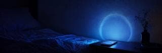 A glowing phone on a nightstand illuminates an empty bed and dried flowers amid scattered items.