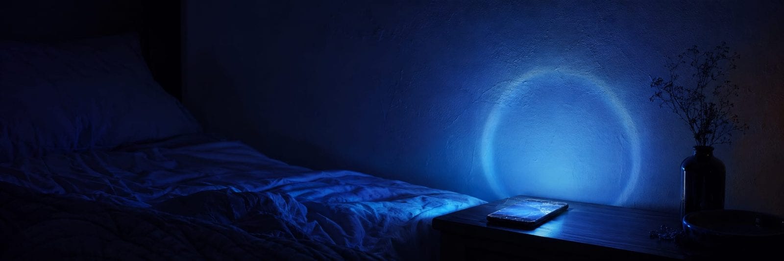 A glowing phone on a nightstand illuminates an empty bed and dried flowers amid scattered items.