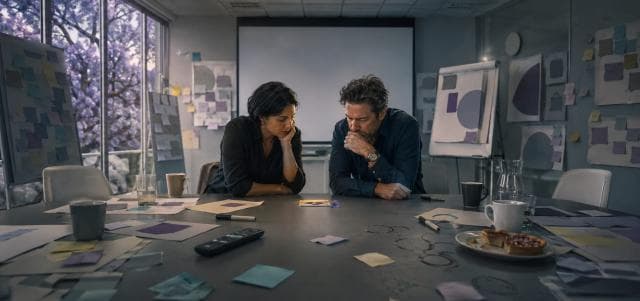 Two people at a cluttered meeting table focus on a small object, hinting at subtle office subversion.