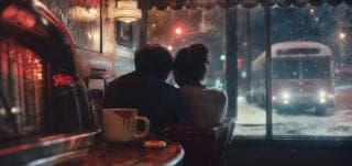 Two people sit close together in a diner at night, watching a snowy street and an approaching bus.