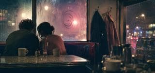 Two people sit together in a diner booth at night, gazing out a window streaked with rain.