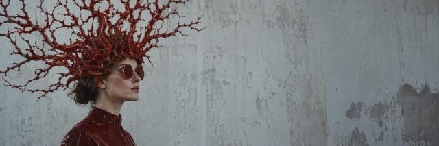 Person in red sunglasses and outfit wearing a branch-like headpiece stands against a textured gray wall.