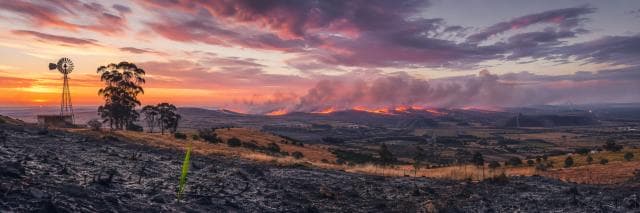 Wildfire burns on distant hills at sunset, with scorched ground, a windmill, and farmland under a dramatic sky.