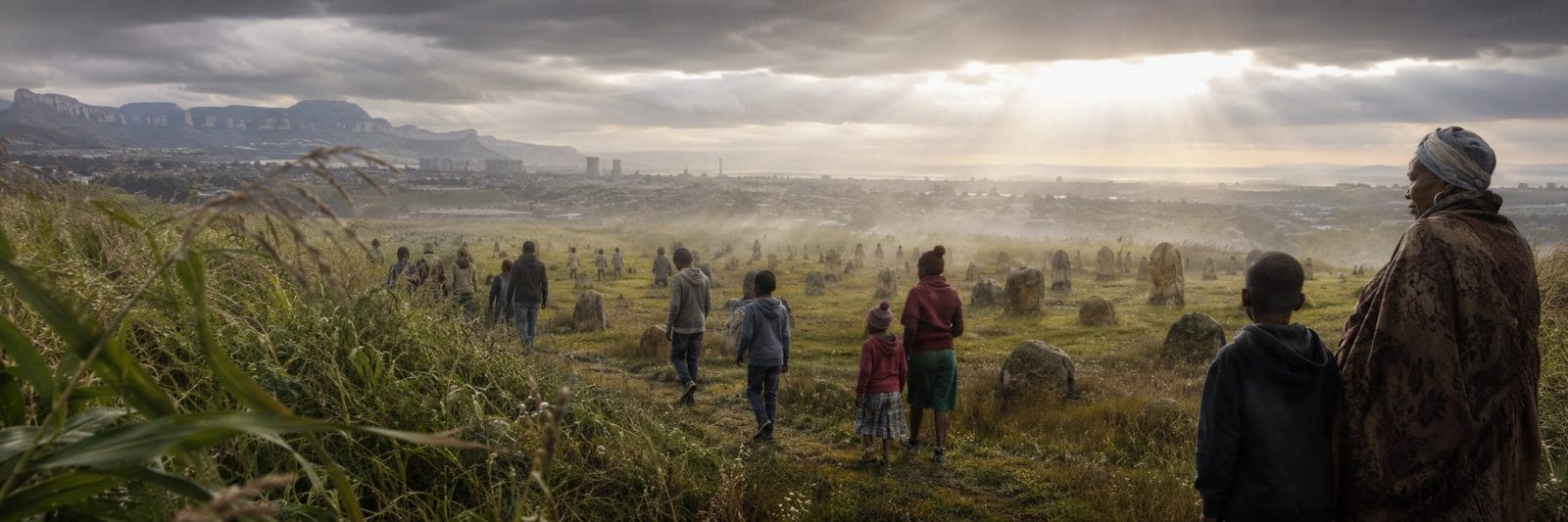 A group walks through a misty field toward a distant city under sun rays breaking through clouds.