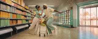 Two people smiling as they push a full shopping cart together down a grocery aisle.