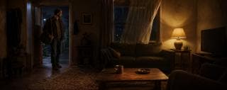 A man with a bag stands in the doorway of a chaotic living room with a sofa, lamp, coffee table, and TV.