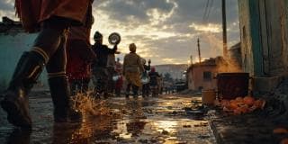 People walk down a wet street at sunset, with metal bowls and steam swirling among food scraps on the ground.