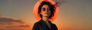 A woman with messy hair stands outdoors at sunset, wearing a swirling robe and a circular red lens that creates a surreal reflection.