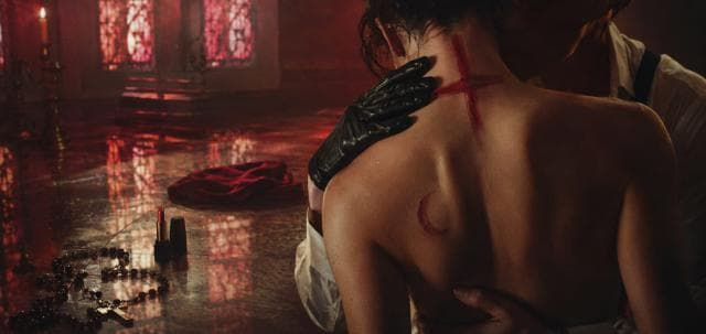 A person with a red cross and crescent is embraced as stained glass, rosary, and lipstick suggest chaos in the room.