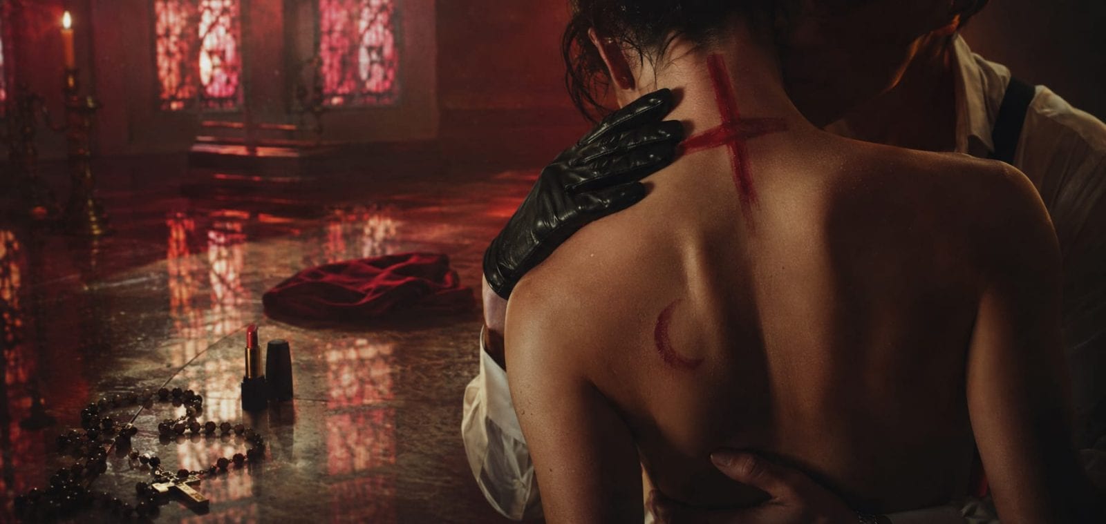 A person with a red cross and crescent is embraced as stained glass, rosary, and lipstick suggest chaos in the room.