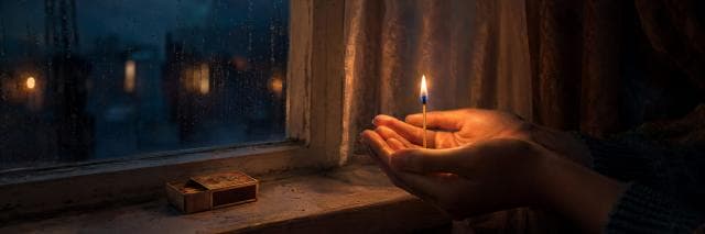 A person shields a lit match near a rainy window, with a matchbox resting quietly on the windowsill.