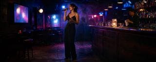 A woman sings into a microphone on stage while a bartender works quietly behind the counter.