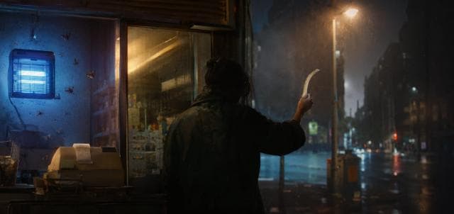 A person stands by a shop counter at night, holding paper, as rain falls on an empty city street.