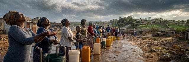 Women wait in line with containers in the rain to collect water in a rural area, with houses visible in the background.