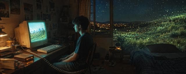 A person at a desk with an old computer looks out at glowing lights in nature, reflecting quietly on the nighttime landscape.