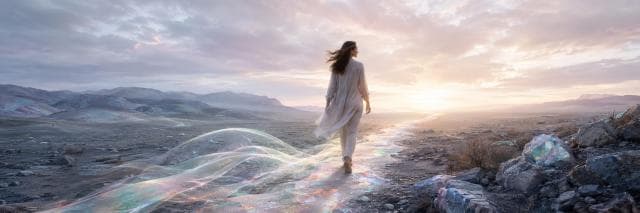 A person in light clothing walks on a glowing iridescent path through rocky terrain at sunrise or sunset, embodying strength and spirit.