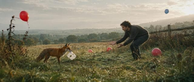 A man crouches in a grassy field, reaching toward a fantastical fox holding a white box with red and blue balloons.