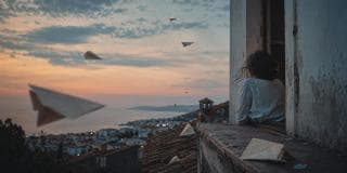 A person sits in a window at sunset, watching paper airplanes fly above a coastal town.