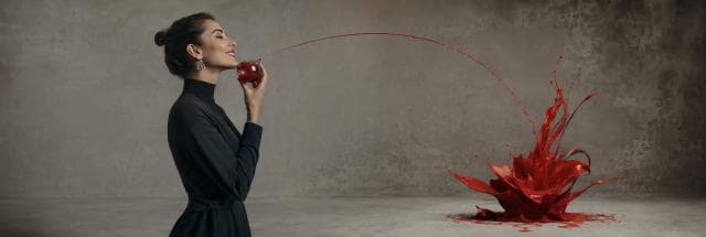 A woman holds a red apple to her chin as red liquid arcs and splashes wildly on the floor.