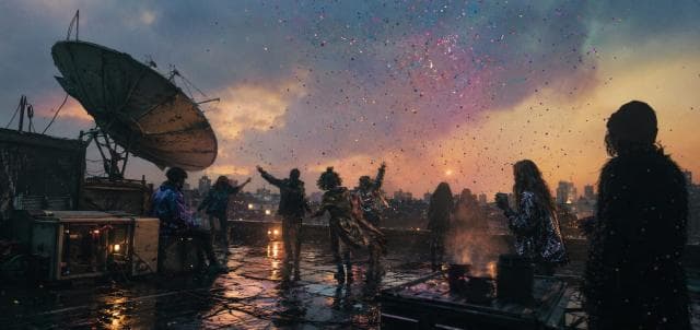 People in shiny outfits celebrate on a rooftop at sunset with confetti, gathered near a satellite dish.