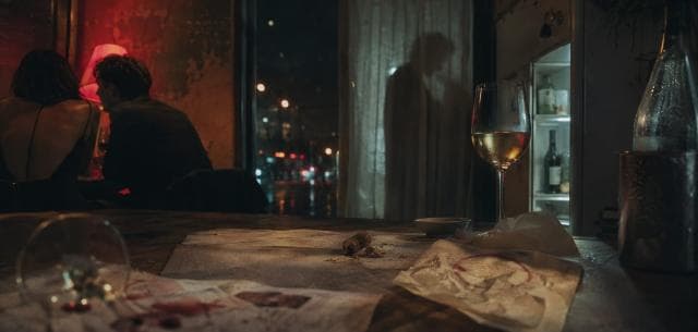A table set with wine glasses and napkins in a restaurant; two people are seated while one stands near an open fridge.