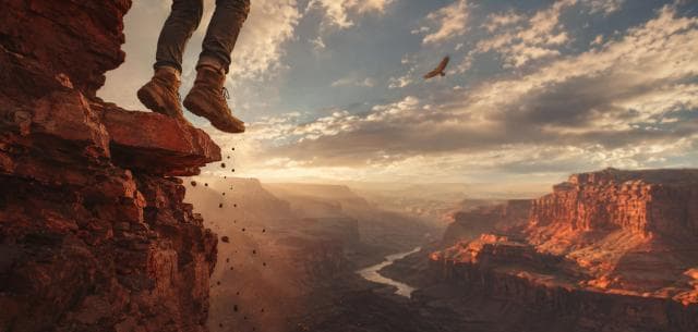 Legs dangle over a rocky cliff at sunset, overlooking a river and canyons, with a bird soaring nearby.