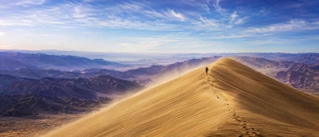A person walks along the ridge of a tall sand dune with mountains and a clear sky in the background.