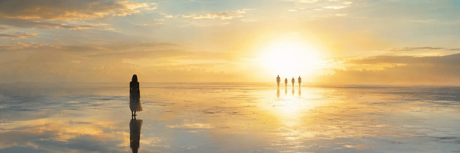 One person stands on a wet reflective surface at sunset while four others walk toward the bright sun.