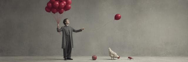 A man stands with red balloons; one is beside a chicken. A red apple and a whimsical red shoe rest on the floor.