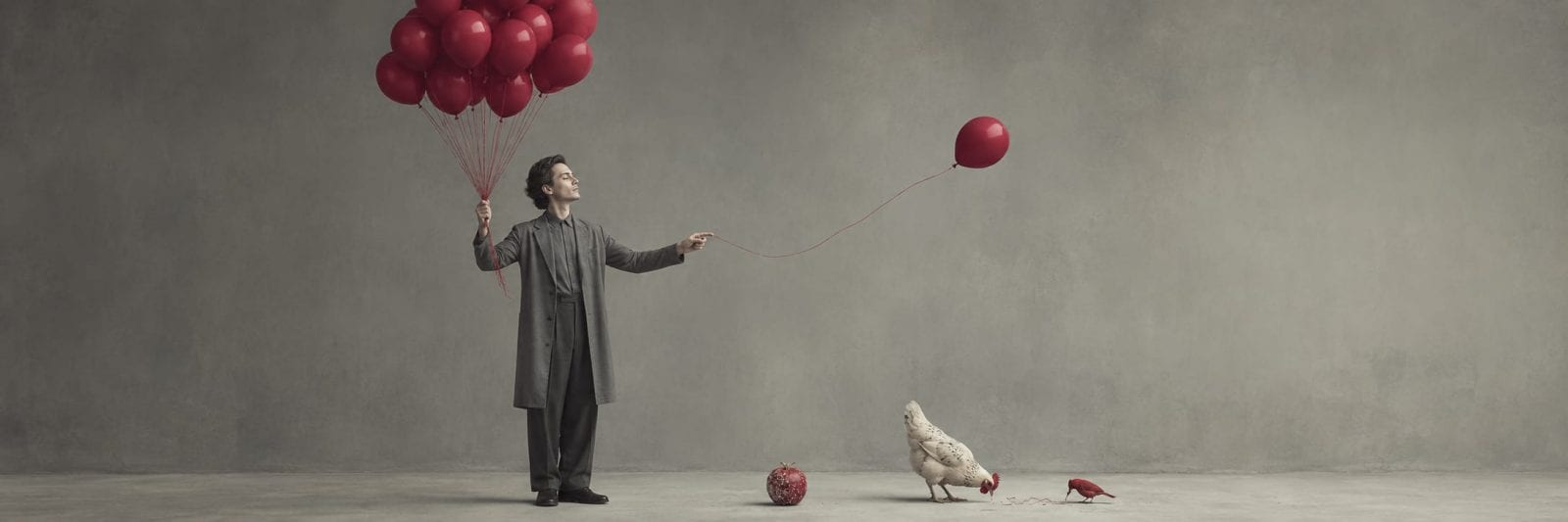 A man stands with red balloons; one is beside a chicken. A red apple and a whimsical red shoe rest on the floor.