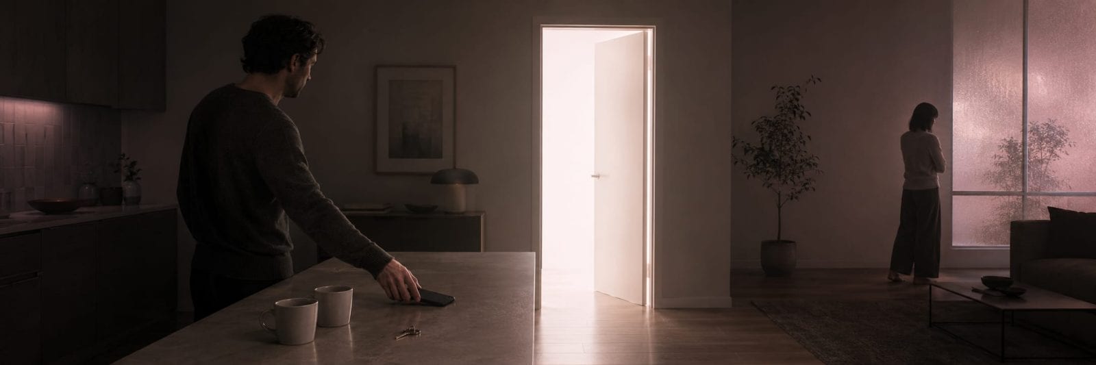 A man holding mugs stands at the kitchen counter while a woman looks outside through an open door.