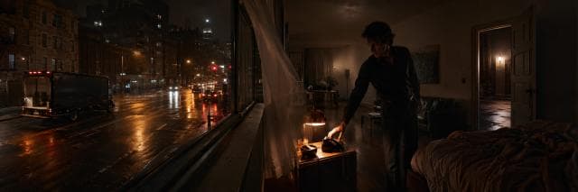 A man in a bedroom answers a rotary phone as rain falls outside and city lights glow, hinting at subtle satire and subversion.