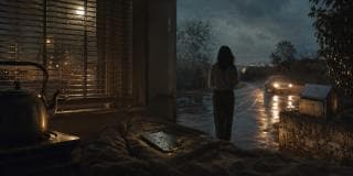 A woman stands confidently on a wet night road as a car approaches, with a lit room and smartphone visible on the bed in front.