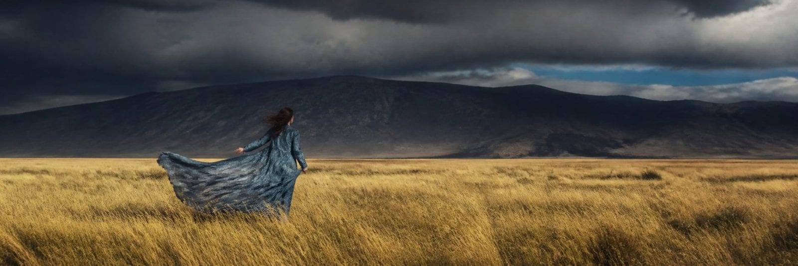 A person in a long blue dress stands in a grassy field with mountains and clouds in the background.