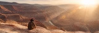 A person with a backpack sits on a rocky ledge, overlooking a canyon river in peaceful reflection.
