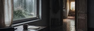 Rain streaks a large window beside a mug and folded paper, while a warm doorway opens into an older house.