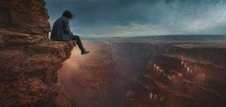 A person sits on a rocky canyon cliff under a surreal, cloudy sky with fantastical cracks in the upper right corner.