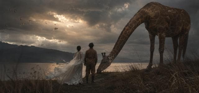 A woman in a white dress and a man reflect by a lake at dusk, with a giraffe grazing nearby under cloudy skies.