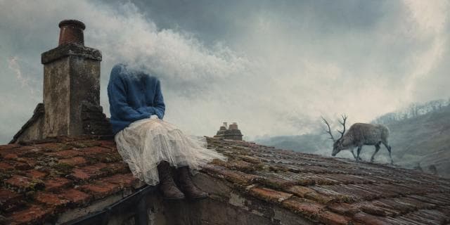 A person with a cloud for a head sits on a tiled roof as a deer walks by on a cloudy, surreal day.