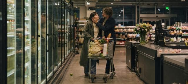 Two people linger by their grocery cart near the checkout in a supermarket at night.