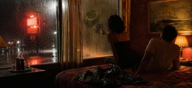 A woman draws on a foggy window as rain falls and a man watches the storm from a motel bed.