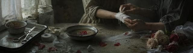 A person wraps another’s bandaged wrist at a table with medical supplies, flowers, and a bowl of water with petals.