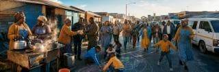 Adults and children gather on a street, cooking or drawing with chalk together among houses and parked vans.